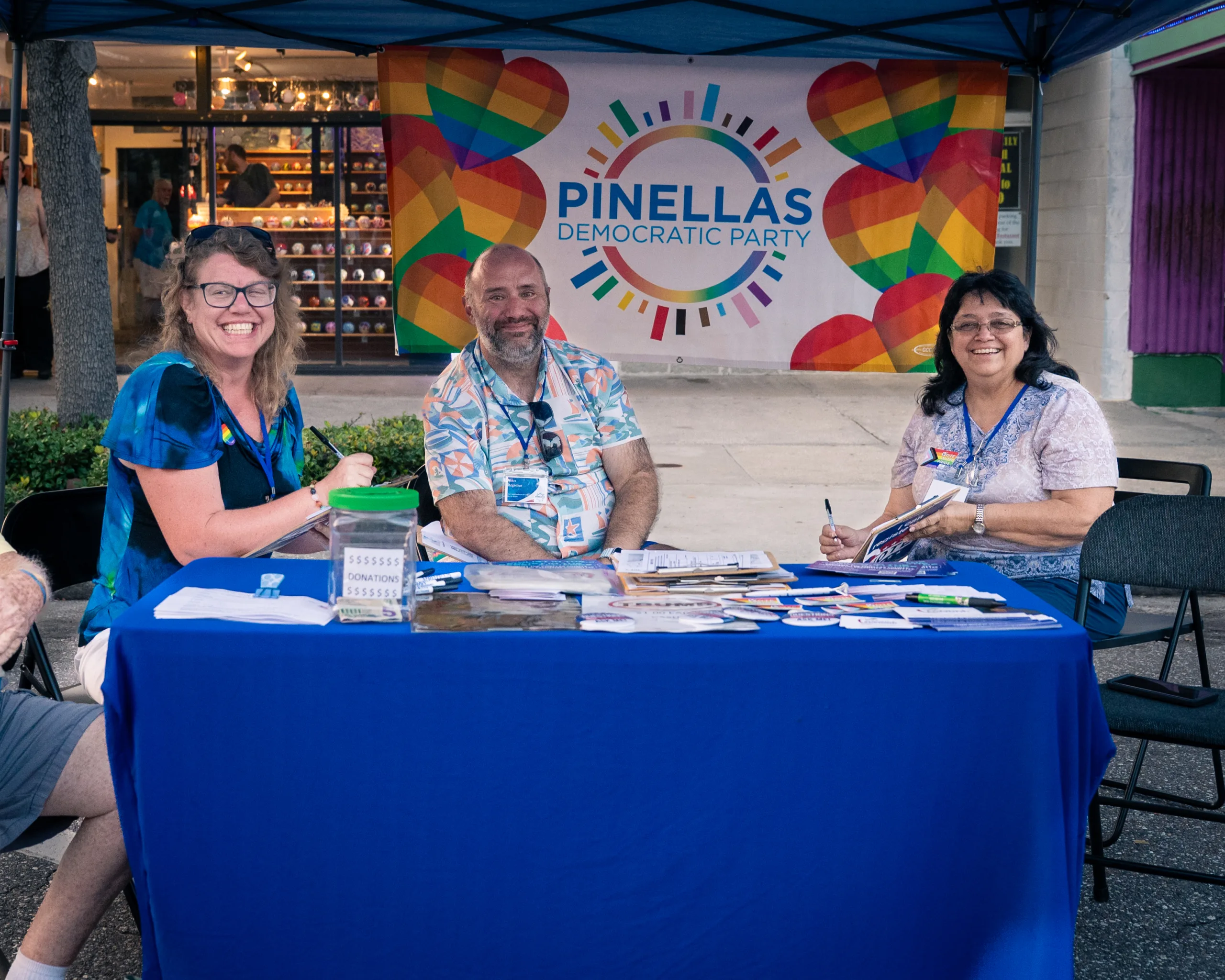 Three people staffing a Pinellas Democrats information table beneath a rainbow party banner.